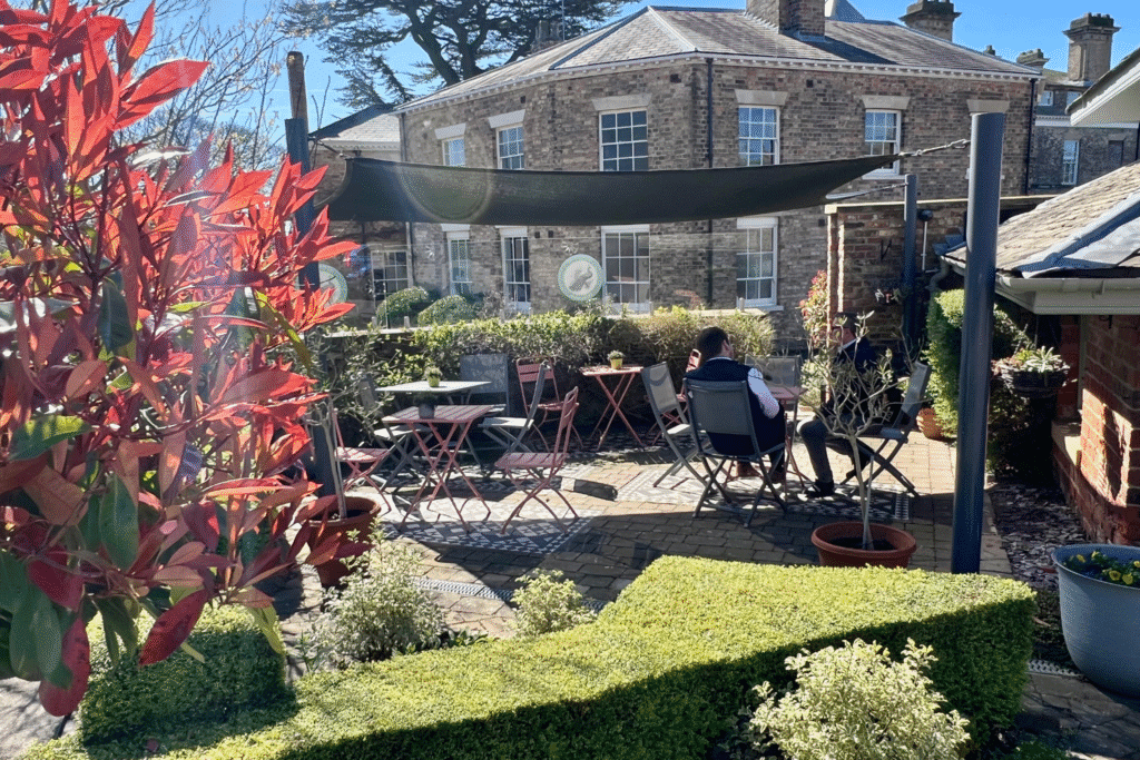 Outdoor seating area of a cafe, covered by a grey sail shade and with shrubs. 2 people sit at a metal bistro table.
