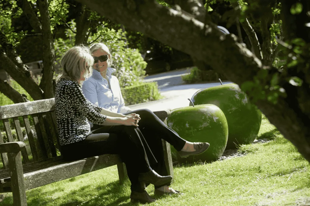 2 females sat on a bench, smiling at one another. The bench sits on grassed parkland, and there are 2 large apple sculptures behind them. Promoting Mental Health Awareness Week