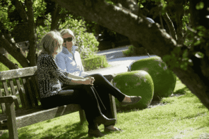 2 females sat on a bench, smiling at one another. The bench sits on grassed parkland, and there are 2 large apple sculptures behind them. Promoting Mental Health Awareness Week