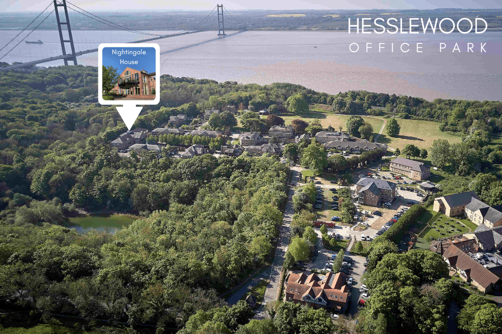 An aerial drone shot of Hesslewood Office Park, a collection of traditional and modern buildings set within a heavily wooded parkland. The buildings are scattered among dense green trees and interconnected by paved roads and parking lots with parked cars visible. A small body of water is partially visible on the left side of the park. In the background, the Humber Bridge spans the wide, muddy brown Humber Estuary, with agricultural fields visible on the opposite bank. The sky is hazy and bright.