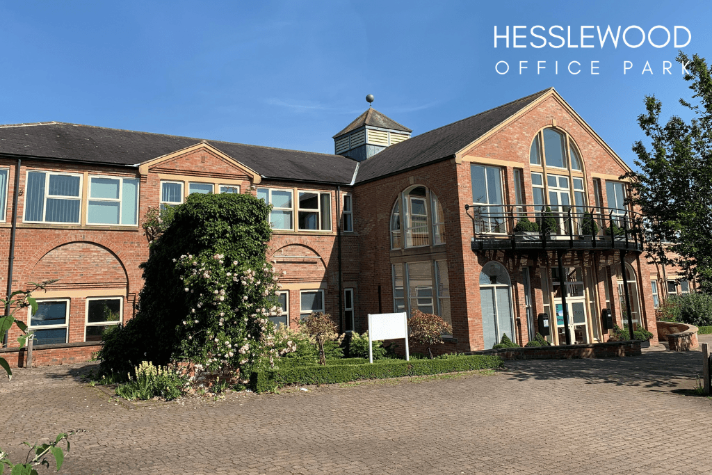 A sunlit, medium-angle shot of a two-story brick office building. The architecture is a mix of styles, with a large, arched window and a small balcony on the right, and more conventional rectangular windows on the left. The building's sign, partially visible in the center, reads "CRODA." A paved brick driveway and landscaped garden beds with a variety of green bushes and flowering plants are in the foreground. A small, overgrown bush with pink flowers is on the left. The sky is clear and blue.