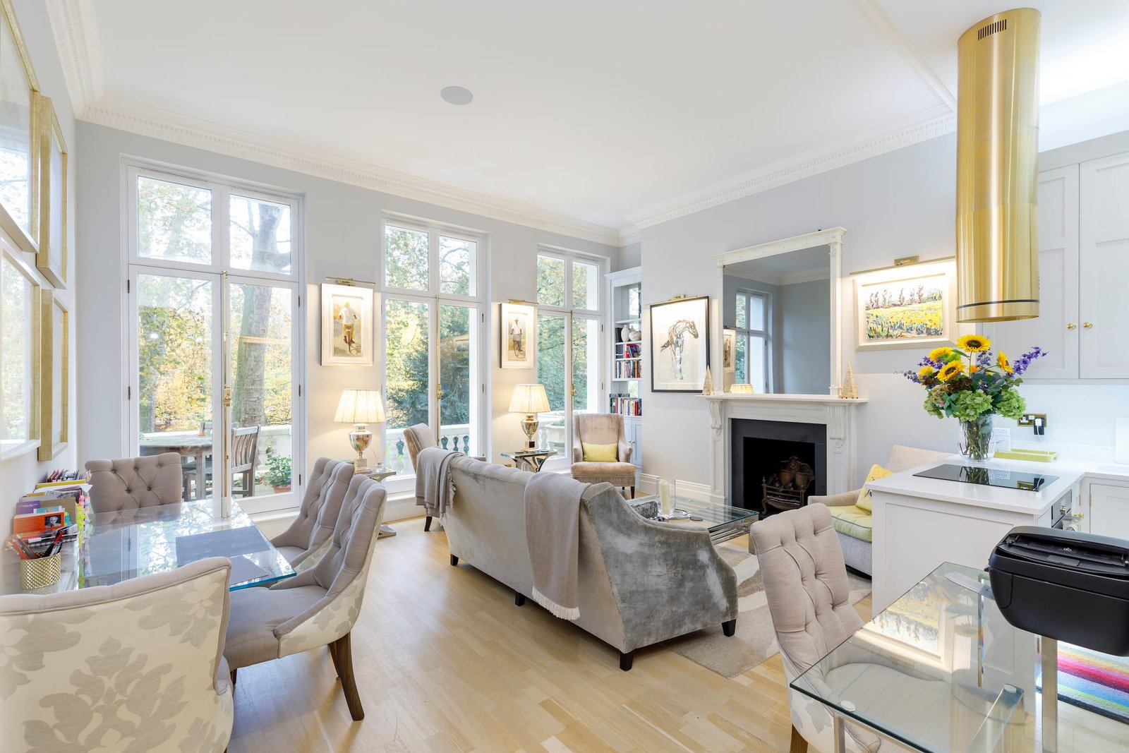 A light-filled open-plan living area with large windows and a glass door opening to an outdoor space. The room is furnished with a mix of light-colored sofas, upholstered chairs, and a glass dining table. A fireplace with a large mirror above it is visible on the right, and the space transitions into a kitchen area with white cabinets and a gold extractor fan.