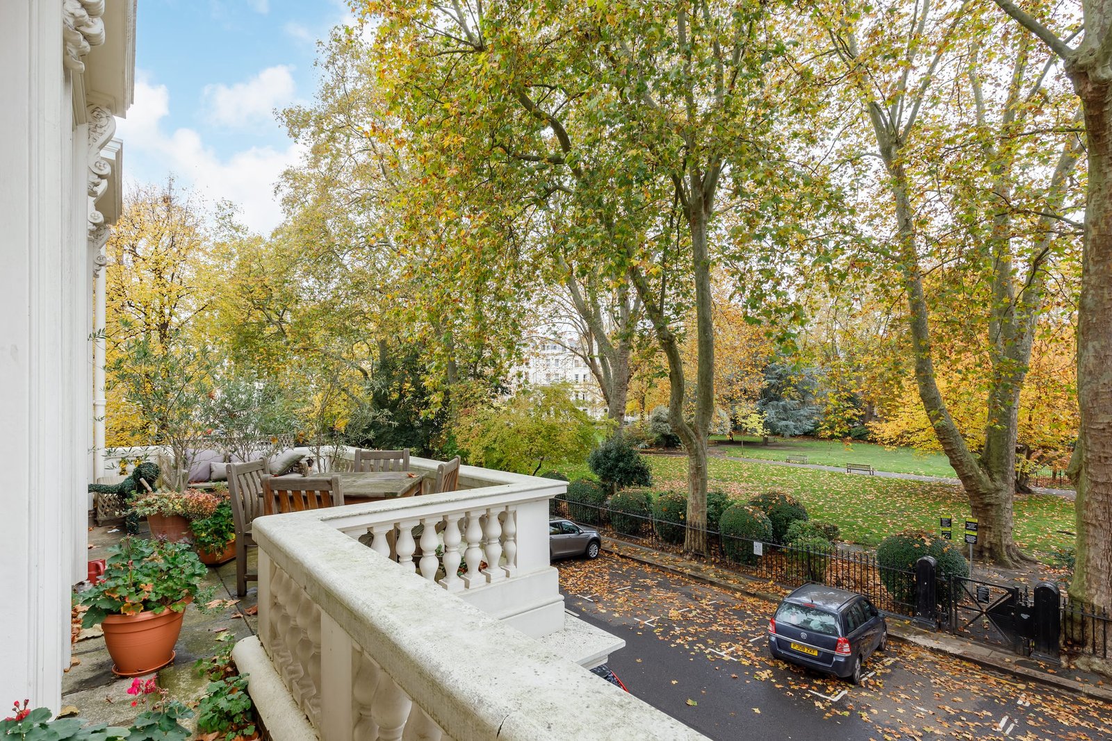 A balcony with a stone balustrade overlooking a leafy street and a park. The trees in the park have yellow and green autumn foliage. A table and chairs are on the balcony, and potted plants are in the foreground.