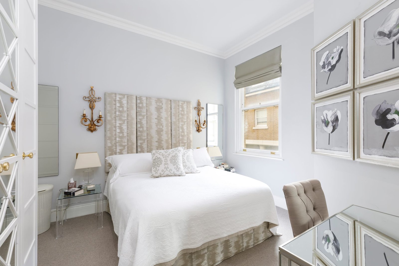 A serene bedroom with light blue walls and a bed with a tall, padded, beige headboard. The bedding is white, and mirrored nightstands are on either side of the bed. A gray tufted chair sits at a mirrored desk. A framed collection of black and white floral prints hangs on the wall to the right.