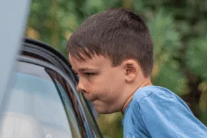 A profile close-up of a young boy with short brown hair, looking down with a serious or focused expression. He is wearing a blue t-shirt and is leaning against what appears to be a black car door. The background is blurred, showing green foliage.