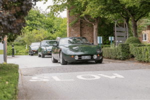 A ground-level shot of three classic cars driving toward the camera. All three cars are a similar shade of dark green. The car in the foreground is a convertible with a curved hood and a British license plate that reads "G5 LVB." The other two cars are visible in a line behind it. The cars are on a paved road with the word "SLOW" painted on the surface in white. A blurred brick building and green trees are visible in the background.