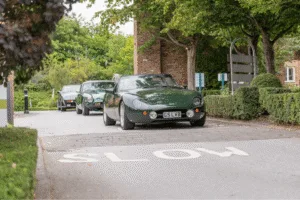 A ground-level shot of three classic cars driving toward the camera. All three cars are a similar shade of dark green. The car in the foreground is a convertible with a curved hood and a British license plate that reads u0022G5 LVB.u0022 The other two cars are visible in a line behind it. The cars are on a paved road with the word u0022SLOWu0022 painted on the surface in white. A blurred brick building and green trees are visible in the background.