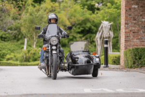 A front-view, medium-angle shot of a man riding a classic chrome motorbike with a black sidecar. The man, who is wearing a black helmet and a black leather jacket, is looking forward. The motorbike has a large windscreen and a round headlight. The sidecar has an open top. A blurred, abstract wooden horse sculpture and green trees are visible in the background.