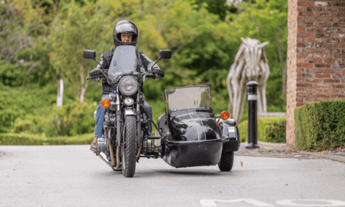 Image from Classic Cars at Hesslewood 2025. A front-view, medium-angle shot of a man riding a classic chrome motorbike with a black sidecar. The man, who is wearing a black helmet and a black leather jacket, is looking forward. The motorbike has a large windscreen and a round headlight. The sidecar has an open top. A blurred, abstract wooden horse sculpture and green trees are visible in the background.