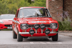A medium close-up, front-view shot of a vintage red race car. The car has white racing stripes, sponsor decals on the side, and a license plate that reads "NTK 555J." Four large circular rally lights with black covers are mounted on the front bumper. The driver is partially visible through the windshield. A blurred brick building is in the background.