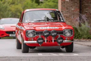 A medium close-up, front-view shot of a vintage red race car. The car has white racing stripes, sponsor decals on the side, and a license plate that reads u0022NTK 555J.u0022 Four large circular rally lights with black covers are mounted on the front bumper. The driver is partially visible through the windshield. A blurred brick building is in the background.