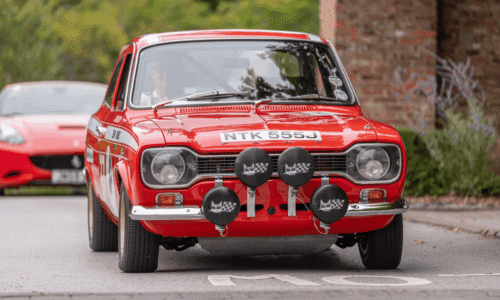 Image from Classic Cars at Hesslewood 2025. A medium close-up, front-view shot of a vintage red race car. The car has white racing stripes, sponsor decals on the side, and a license plate that reads "NTK 555J." Four large circular rally lights with black covers are mounted on the front bumper. The driver is partially visible through the windshield. A blurred brick building is in the background.