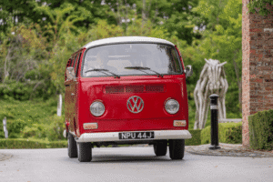 A low-angle, front-view shot of a vintage red and white Volkswagen camper van. The van has a license plate that reads "NPU 44J." The driver is visible through the windshield. A blurred, abstract wooden horse sculpture and a lamp post are visible in the background, surrounded by green trees.