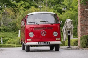 A low-angle, front-view shot of a vintage red and white Volkswagen camper van. The van has a license plate that reads u0022NPU 44J.u0022 The driver is visible through the windshield. A blurred, abstract wooden horse sculpture and a lamp post are visible in the background, surrounded by green trees.