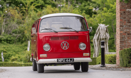 Image from Classic Cars at Hesslewood 2025. A low-angle, front-view shot of a vintage red and white Volkswagen camper van. The van has a license plate that reads "NPU 44J." The driver is visible through the windshield. A blurred, abstract wooden horse sculpture and a lamp post are visible in the background, surrounded by green trees.