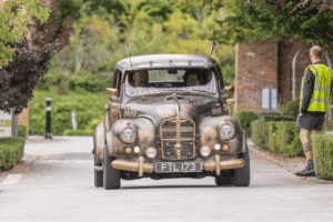 A low-angle shot of a custom-built car with a steampunk or post-apocalyptic aesthetic. The car, which has a matte black finish with bronze or gold detailing, appears to be an older model with circular headlights. It features various decorative elements, including skulls on the grille and horns on the roof. Two people are visible in the front seats. The car is driving down a paved road surrounded by green trees and hedges.