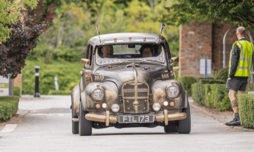 Image from Classic Cars at Hesslewood 2025. A low-angle shot of a custom-built car with a steampunk or post-apocalyptic aesthetic. The car, which has a matte black finish with bronze or gold detailing, appears to be an older model with circular headlights. It features various decorative elements, including skulls on the grille and horns on the roof. Two people are visible in the front seats. The car is driving down a paved road surrounded by green trees and hedges.