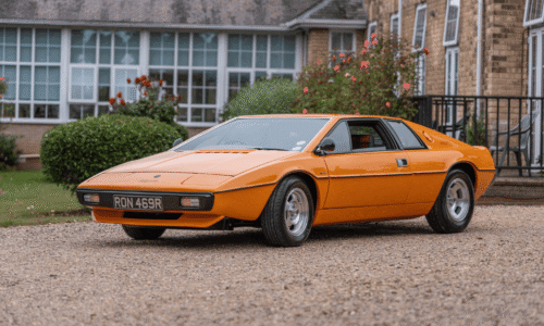 Image from Classic Cars at Hesslewood 2025. A profile, eye-level shot of a classic, orange Lotus Esprit sports car. The car is parked on a gravel driveway in front of a large brick building with many windows and blooming rose bushes. The car's license plate reads "RON 469R."