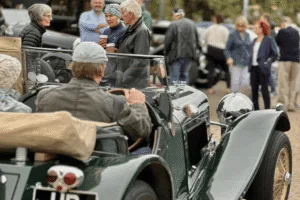 A crowd of people gathered around vintage cars at an outdoor event. In the foreground, a person in a flat cap is seated in a classic green convertible. In the background, other people are visible, including a person holding two disposable coffee cups, talking to another person.