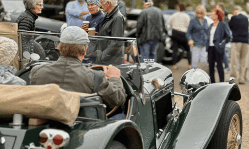 Image from Classic Cars at Hesslewood 2025. A crowd of people gathered around vintage cars at an outdoor event. In the foreground, a person in a flat cap is seated in a classic green convertible. In the background, other people are visible, including a person holding two disposable coffee cups, talking to another person.