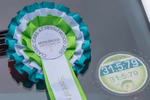 A close-up of a large rosette award, with pleated ribbons in shades of white, light green, and teal. The circular centre of the rosette has text around a tree logo that reads u0022CLASSIC CARS AT HESSLEWOOD 2025u0022 and u0022HESSLEWOOD CHARITABLE CAUSES FUND.u0022 A long tail of a green ribbon with white stripes hangs down from the rosette. To the right, a circular British car tax disc is visible on a glass surface.