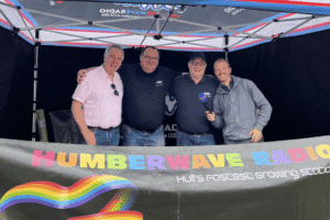 A group of four men are standing under a tent with a Humberwave Radio sign. The man on the far right is holding a microphone with the radio station's logo and is smiling. The men are dressed casually in t-shirts and jeans. A banner in the foreground features a rainbow and the words "HUMBERWAVE RADIO"