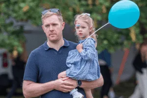 A candid, medium close-up shot of a person holding a young child. The person is looking slightly to the left with a serious expression, wearing a navy polo shirt and sunglasses pushed up on their head. The child is holding a light blue balloon and has a face painting design on their cheek. The child is wearing a patterned blue dress.