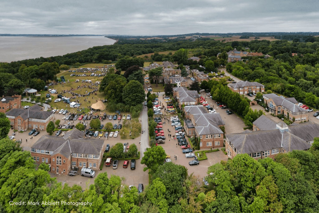 An aerial drone shot of Hesslewood Office Park during the 2025 Classic Cars at Hesslewood event. The image shows a large collection of classic cars filling the parking lots and grounds, surrounded by brick buildings and green trees. People are visible mingling around the cars and in a large open field on the left. The field also features white tents, indicating a larger event. A large body of water, the Humber Estuary, is visible in the background, with the land on the far bank in the distance. The sky is grey and overcast. A small caption in the bottom left corner reads, "Credit: Mark Abblett Photography."