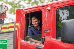 A man in a navy fire officers uniform, with a serious yet slightly amused expression, is seated in the passenger side of a red fire truck. He is looking at the camera, with the window rolled down. The side of the fire truck is visible, with the words "CrewCab" and "SPARE" on the door. The background is a mix of blurred greenery and a light-colored sky.