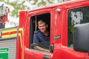 A man in a navy fire officers uniform, with a serious yet slightly amused expression, is seated in the passenger side of a red fire truck. He is looking at the camera, with the window rolled down. The side of the fire truck is visible, with the words u0022CrewCabu0022 and u0022SPAREu0022 on the door. The background is a mix of blurred greenery and a light-colored sky.