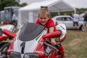 A smiling child sits on a red and white motorbike with the number u00221u0022 on the front fairing. The child is wearing a red and green jersey and is gripping the handlebars. A white racing helmet rests on the seat behind them. A white tent and a car are visible in the background.