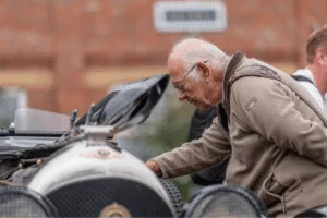 A profile close-up of a person with a bald head and a white mustache, wearing glasses. The person is looking down intently at the front of a vintage car, which has a shiny silver grill and a black hood. They are wearing a beige fleece jacket over a blue shirt. The background is a blurred brick building and green trees.