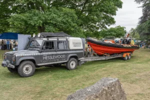 A Land Rover Defender marked 'Humber Rescue' towing a large orange and black rescue boat on a trailer, parked on grass at an outdoor event with trees and people in the backgro