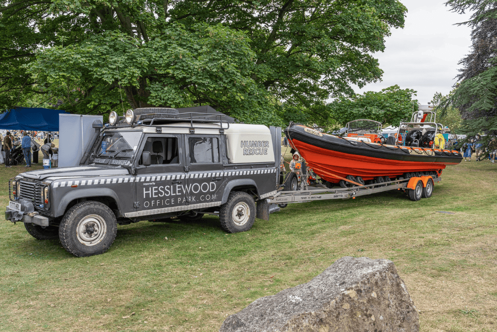 A Land Rover Defender marked 'Humber Rescue' towing a large orange and black rescue boat on a trailer, parked on grass at an outdoor event with trees and people in the backgro