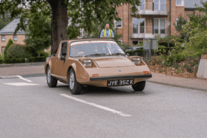 A head-on, low-angle shot of a 'Clan' vehicle driving on a paved road. The vehicle has a light brown, wedge-shaped body, with large, square headlights and a windshield. A person is visible in the driver's seat. In the background, a person in a bright yellow vest is standing on the sidewalk. A building with brick and windows is also visible behind a tree.