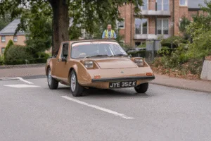 A head-on, low-angle shot of a 'Clan' vehicle driving on a paved road. The vehicle has a light brown, wedge-shaped body, with large, square headlights and a windshield. A person is visible in the driver's seat. In the background, a person in a bright yellow vest is standing on the sidewalk. A building with brick and windows is also visible behind a tree.