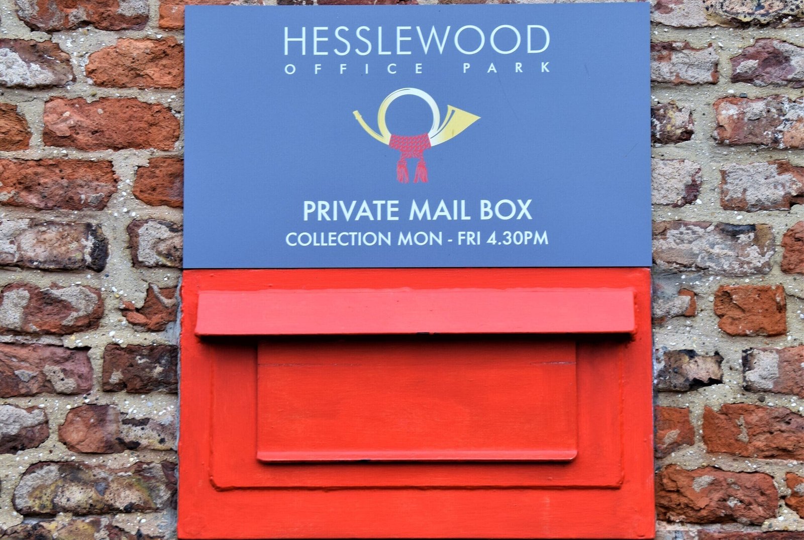 A close-up of a red private mail box mounted on a brick wall. A blue sign above the mailbox features the words "HESSLEWOOD OFFICE PARK," a logo of a golden bugle with a red ribbon, and the text "PRIVATE MAIL BOX" with "COLLECTION MON - FRI 4.30PM" below it. 