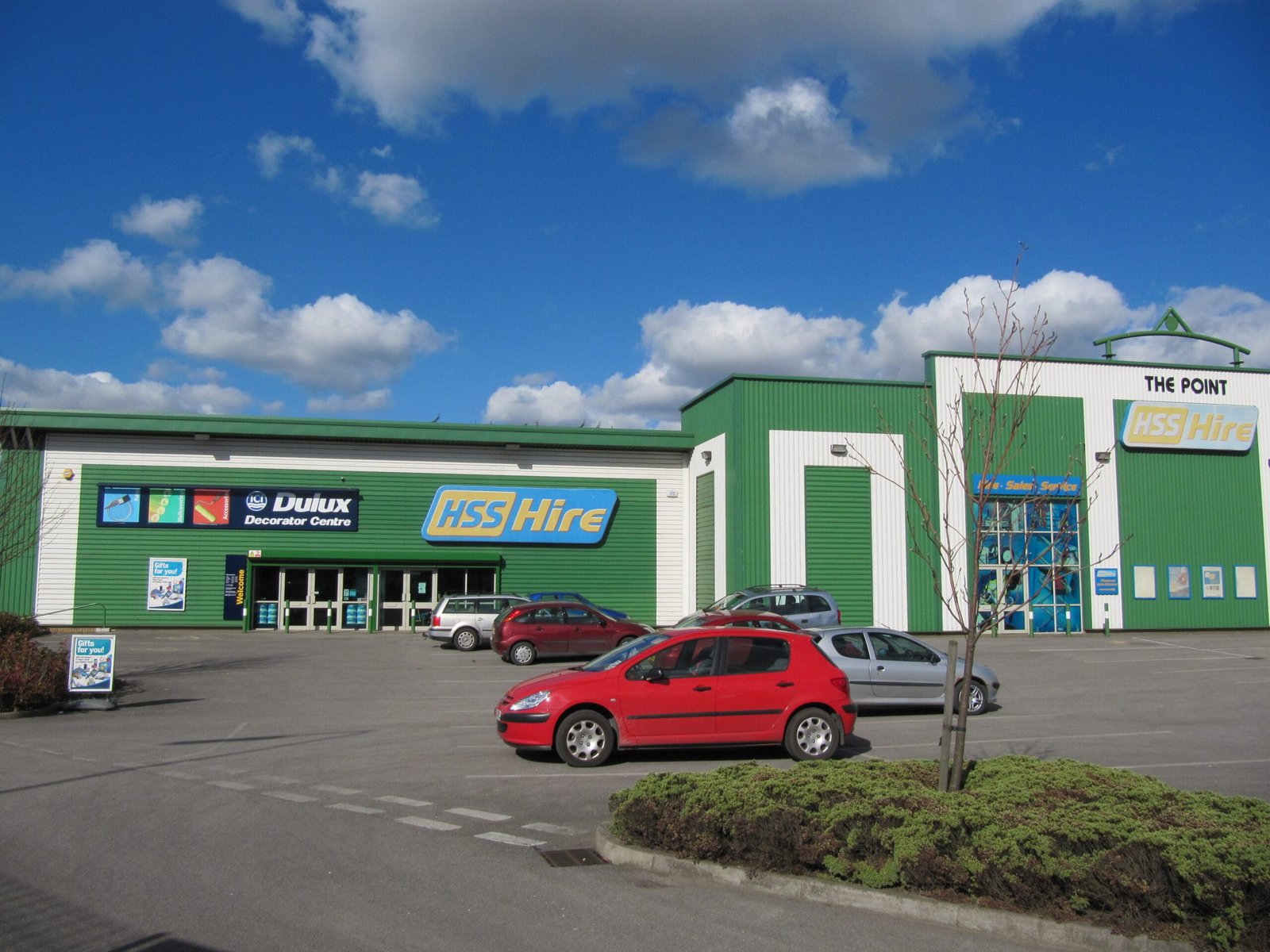 A medium-angle shot of a long, green commercial building with white vertical stripes. The building, named "The Point," houses three businesses: an HSS Hire tool rental store in the center, flanked by Dulux  location to the left and a Bathstore to the right. A paved parking lot sits in front of the building, with a few cars parked in front of each business. A well-maintained garden bed with a low, green hedge and a small tree separates the building from the parking lot. The sky is partly cloudy.