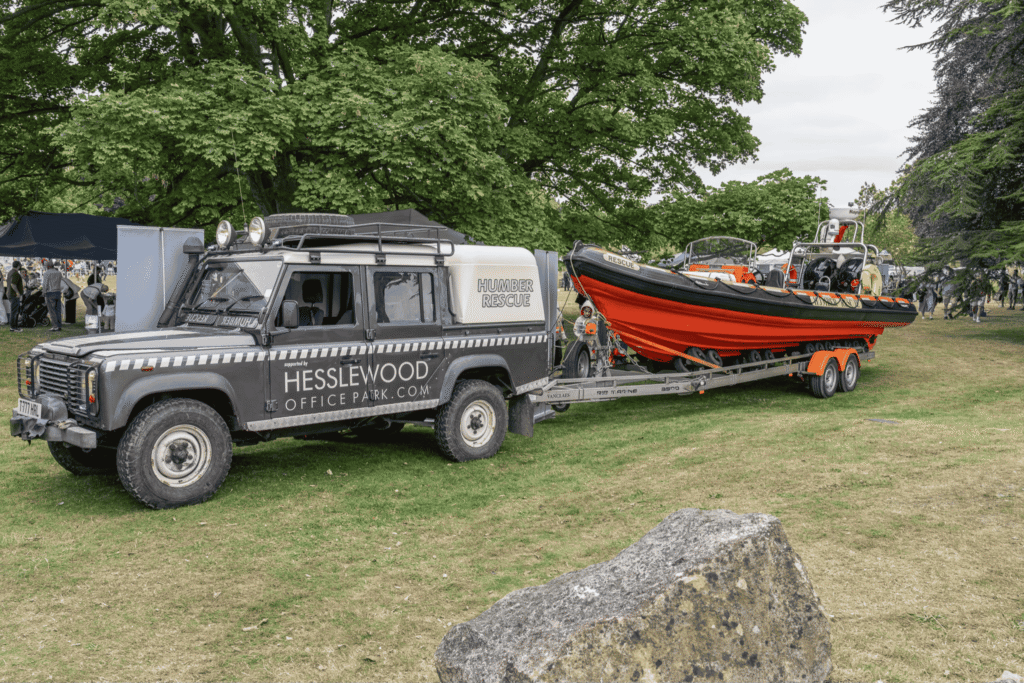 A Humber Rescue vehicle towing a bright orange and black lifeboat into Classic Cars at Hesslewood 2025, displayed on the grounds beneath large trees.