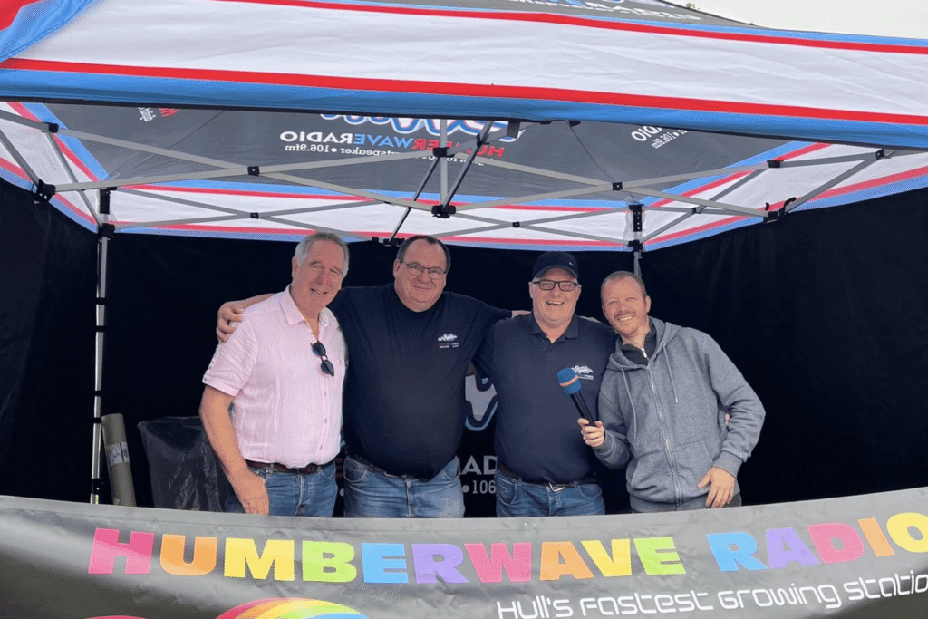 The Humber Wave Radio team smiling together under their event tent at Classic Cars at Hesslewood 2025, with a banner promoting the station displayed at the front