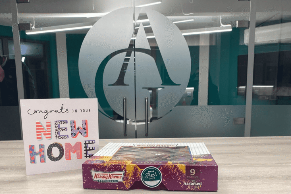 Gift card reading ‘Congrats on your new home’ and a box of Krispy Kreme doughnuts placed on a table in front of frosted glass office doors featuring a large circular logo (initials of Adams and Green)