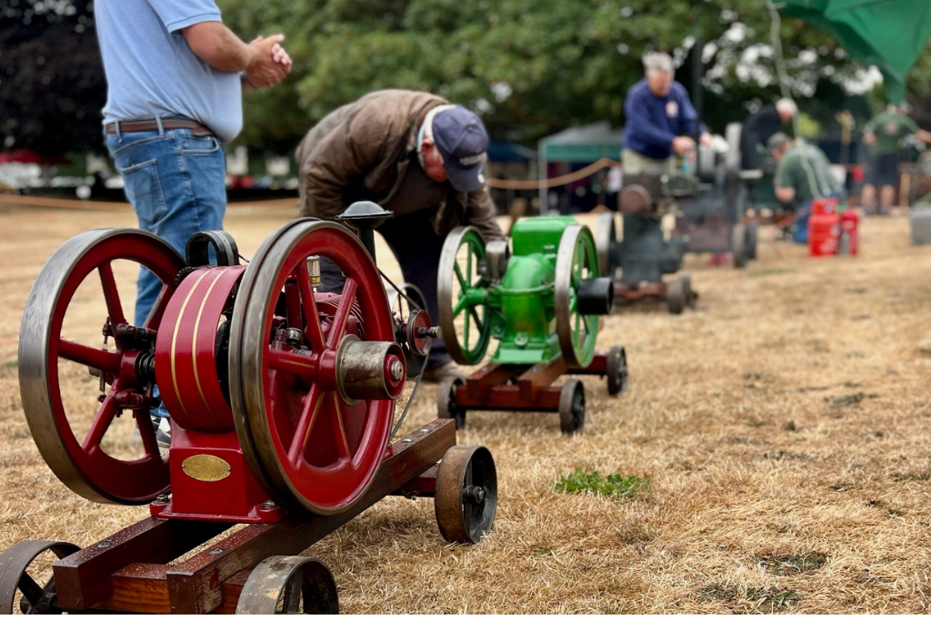 A row of stationary vintage engines. One bystander looks on, and one male hunches over the middle one in the row