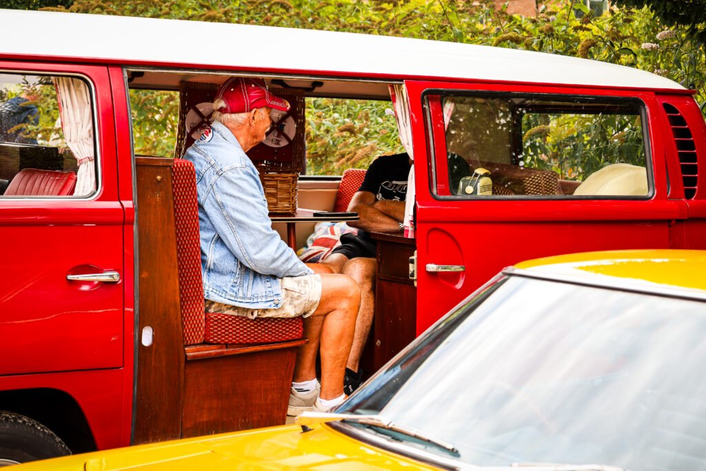 Close up of male wearing cap, sat opposite another person, inside a red VW Campervan