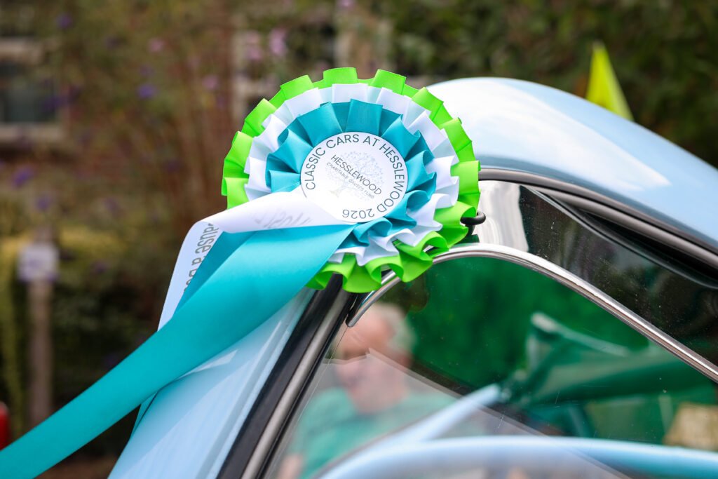 Close up of a an award rosette in green, white and blue, that reads Classic Cars at Hesslewood 2026. attached to the bonnet of a pale blue vehicle.